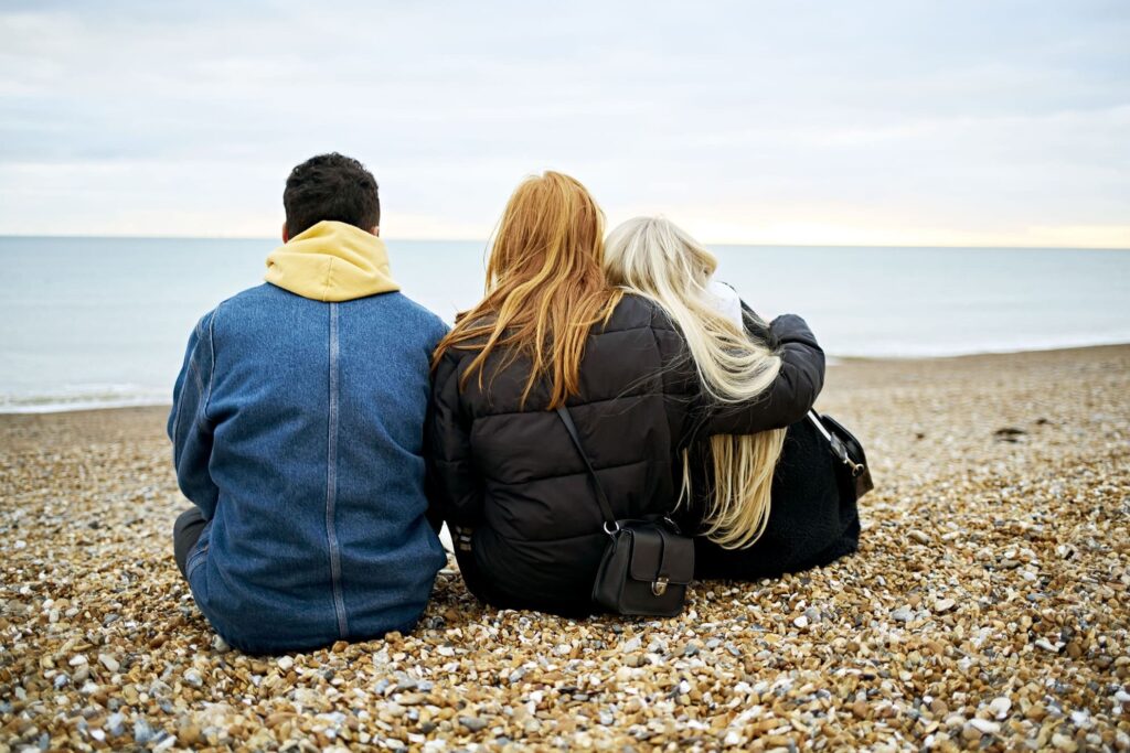 Three young people sitting on beach with their backs to us.