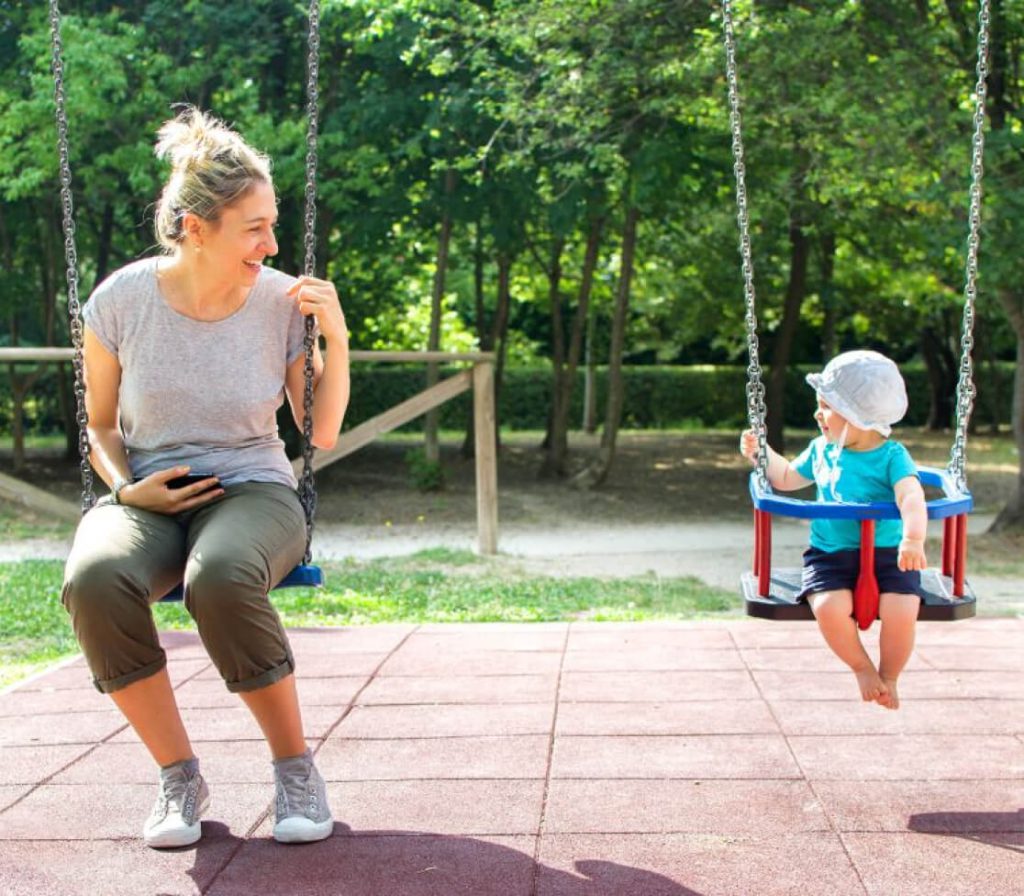 A mother and child each sit on a swing whilst facing towards each other and smiling.