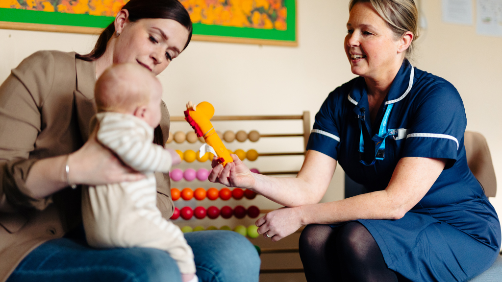 A health visitor holding up a toy in front of a young baby, who is being held by its mother.
